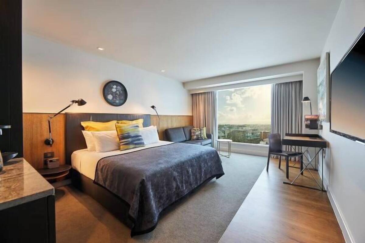 A modern hotel room with a large bed, a desk, and a view of the city.