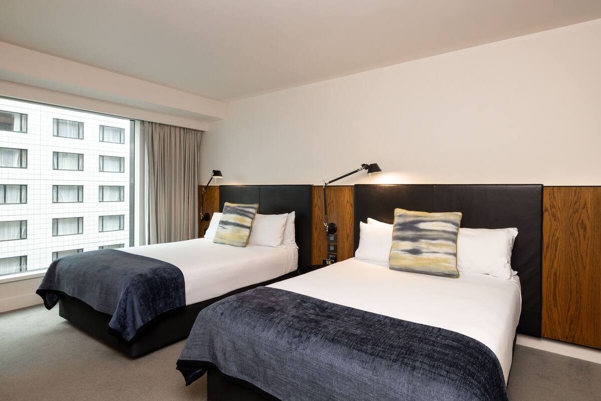 A hotel room with two beds, each with a headboard, a window with curtains, and a view of a building.