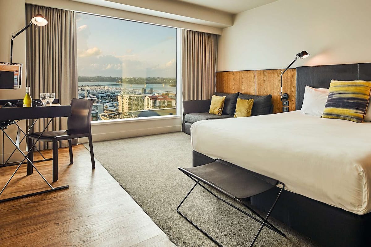 A hotel room with a large bed, a desk, and a chair, with a view of a cityscape through large windows.