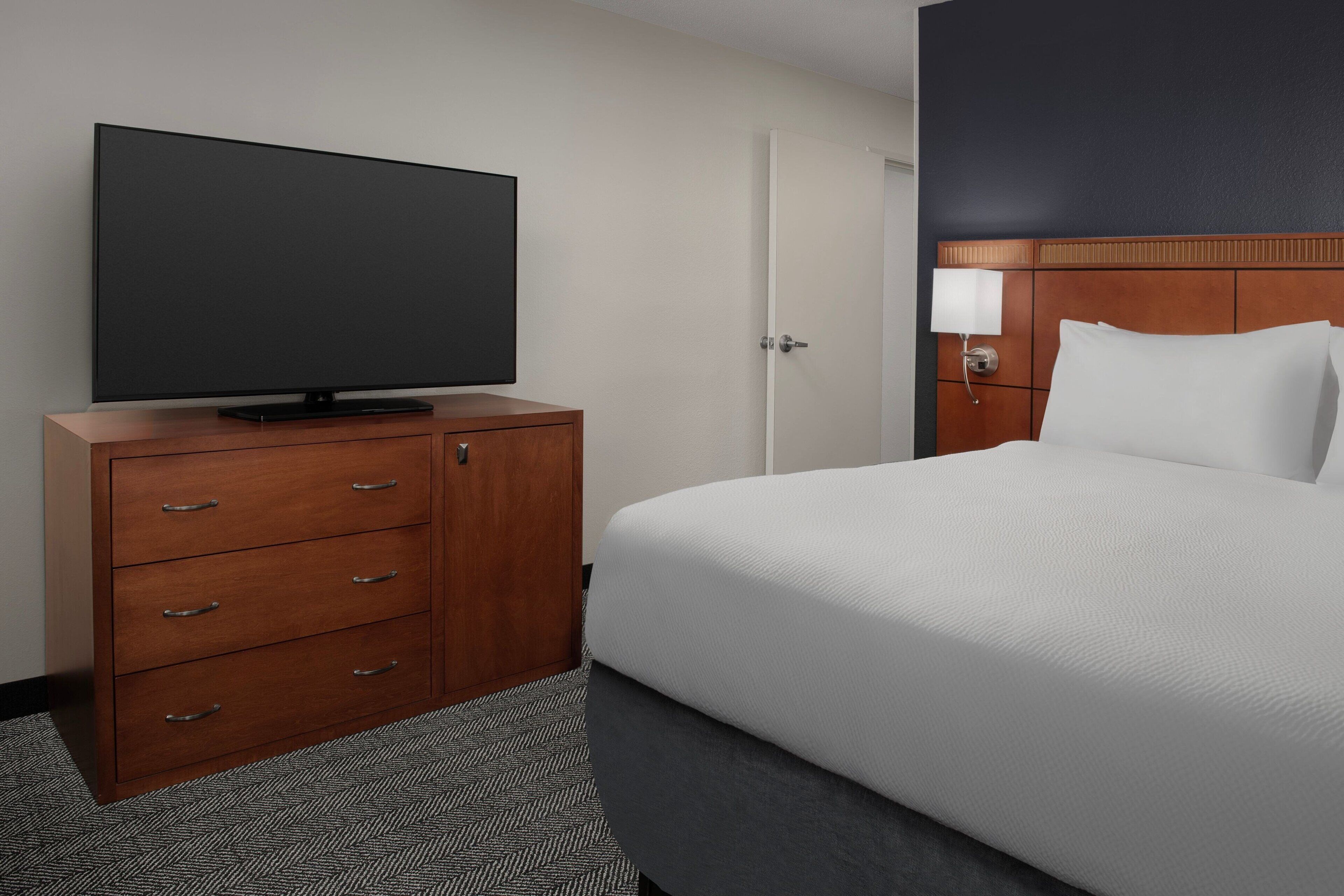 Quarto de hotel com televisão de tela plana sobre um móvel de madeira, cama com roupa de cama branca e porta com design em painel de madeira.