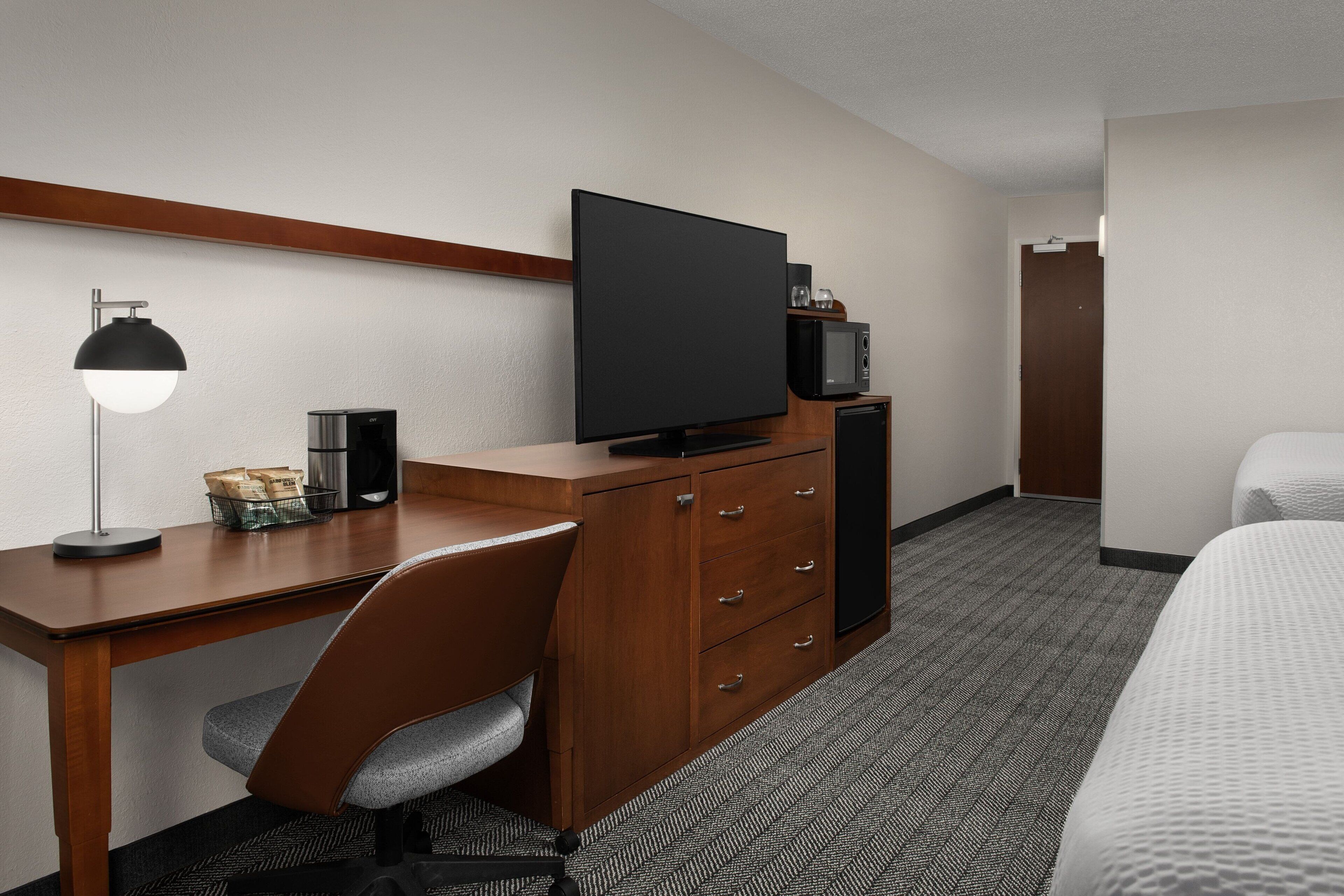 A hotel room with a wooden desk, a flat-screen TV, a chair, a bed, and a microwave.
