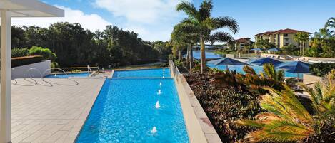 Una piscina con un elemento acuático de agua cristalina y azul, rodeada de palmeras y vista a un edificio de resort.