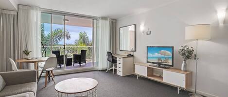 Une chambre d’hôtel avec un canapé, une table ronde, un bureau avec une chaise, une télévision, une lampe et une grande fenêtre avec des rideaux.