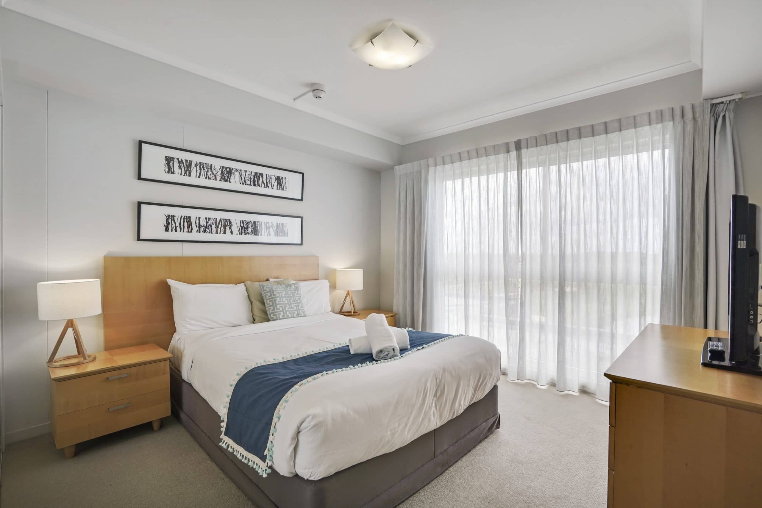 A hotel room with a bed, two bedside tables, a desk, and a large window with curtains.