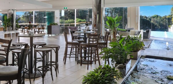A spacious indoor area with multiple tables and chairs, a large window overlooking a pool, and several potted plants. - Pelican Waters Resort (Pelican Waters)