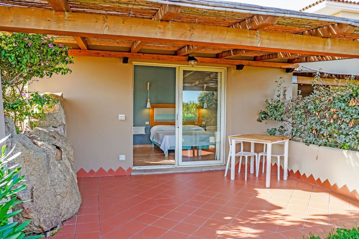 A covered patio with a bed, a table, and chairs, surrounded by plants and a rock.