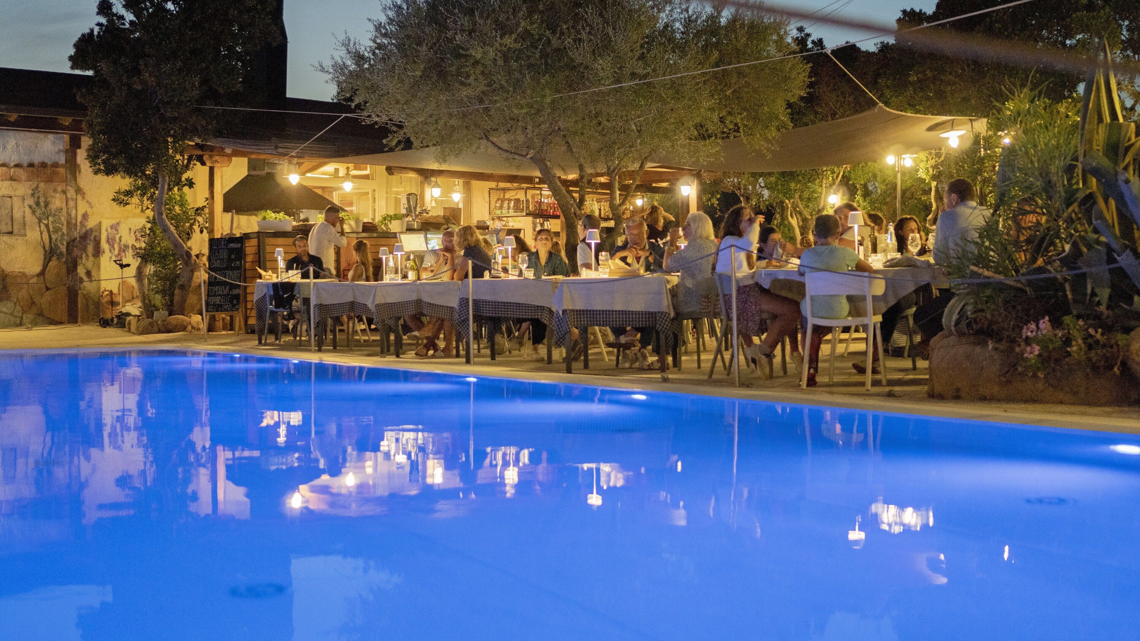 An outdoor dining area with tables and chairs, a pool, and a building with a bar in the background.