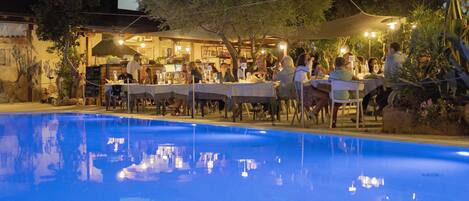 An outdoor dining area with tables and chairs, a pool, and a building with a bar in the background.