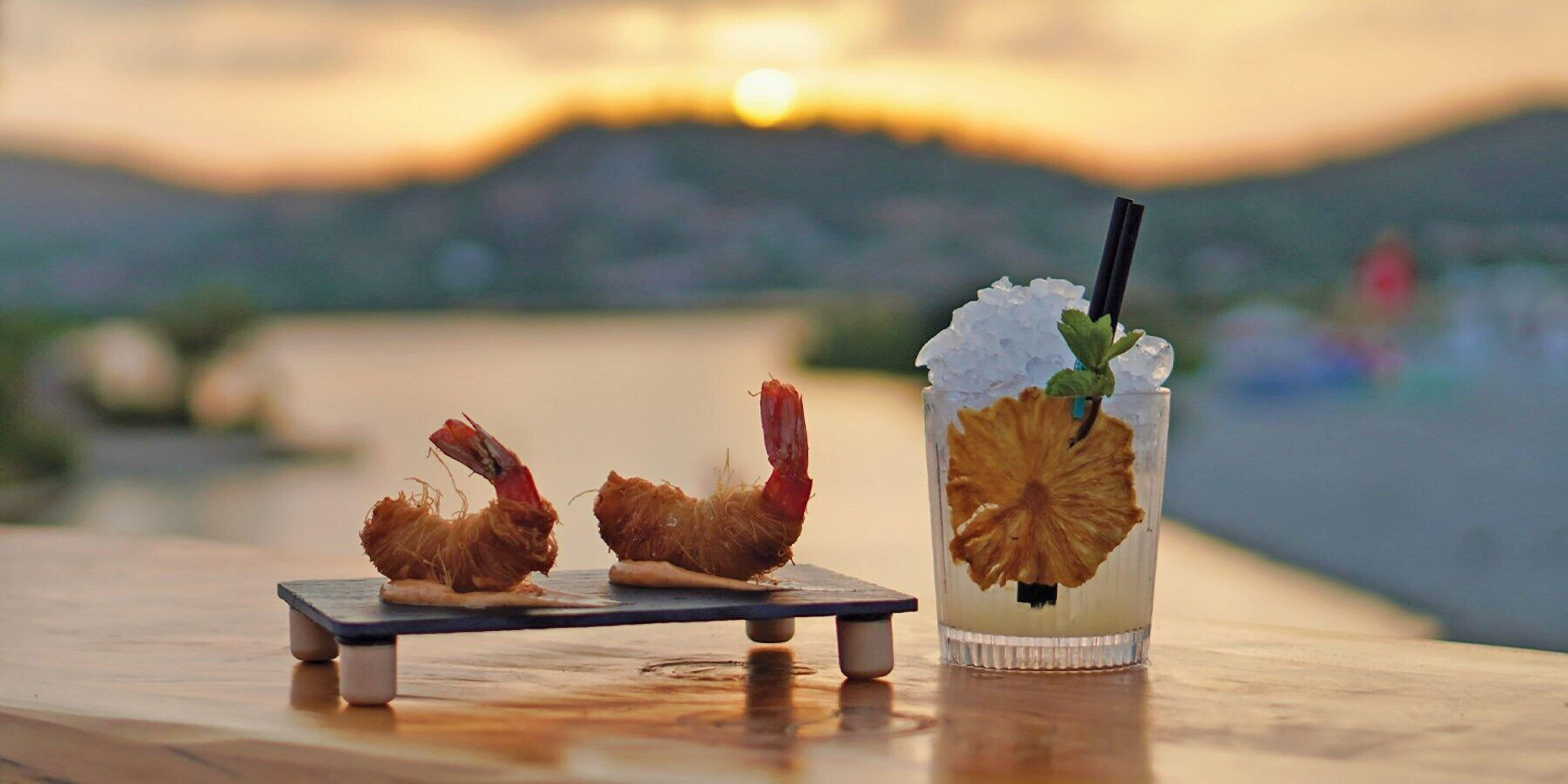 A sunset view with a table featuring two shrimp appetizers and a cocktail with a dried fruit garnish.