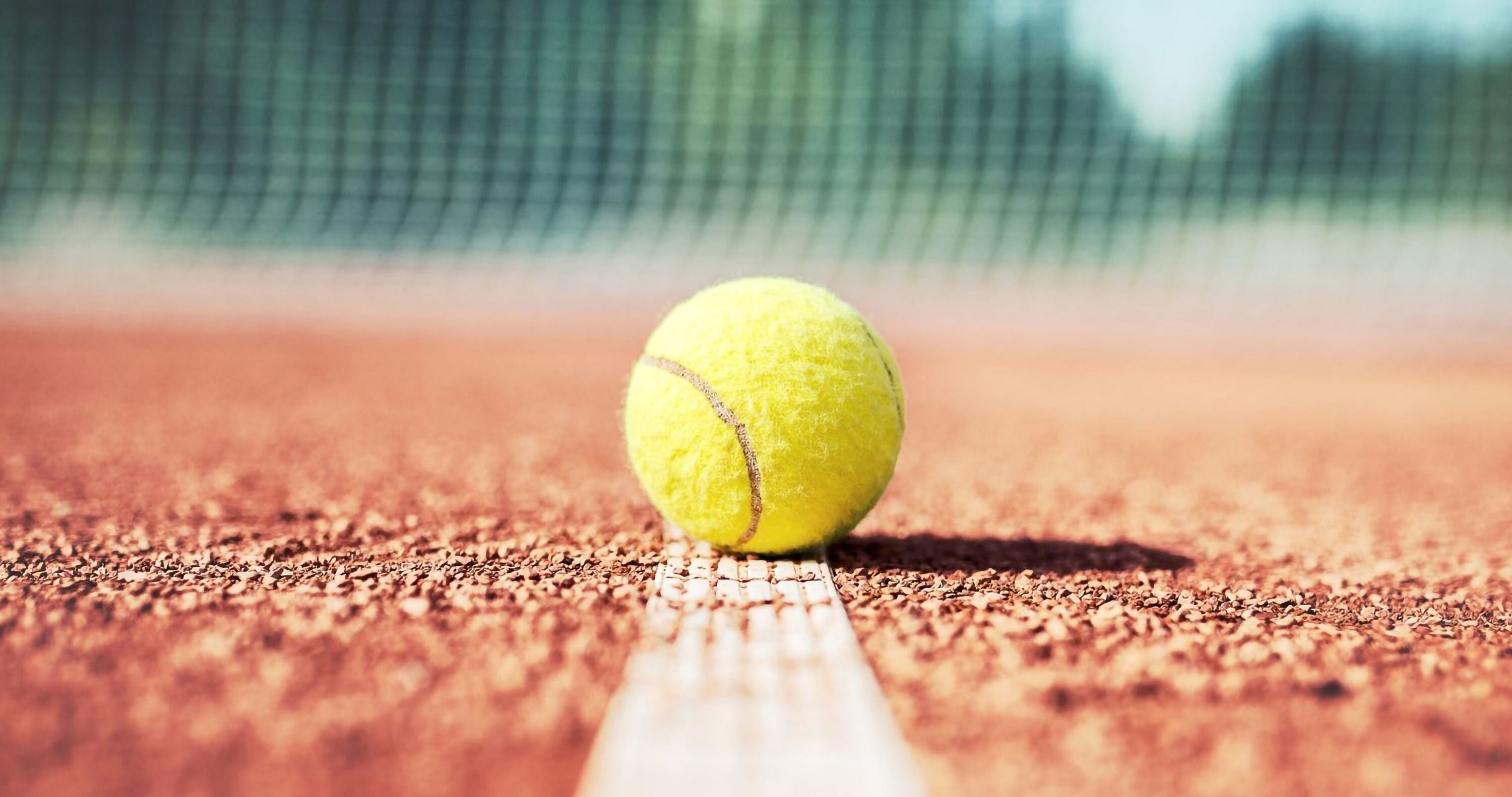 A tennis ball on a clay court with a net in the background.
