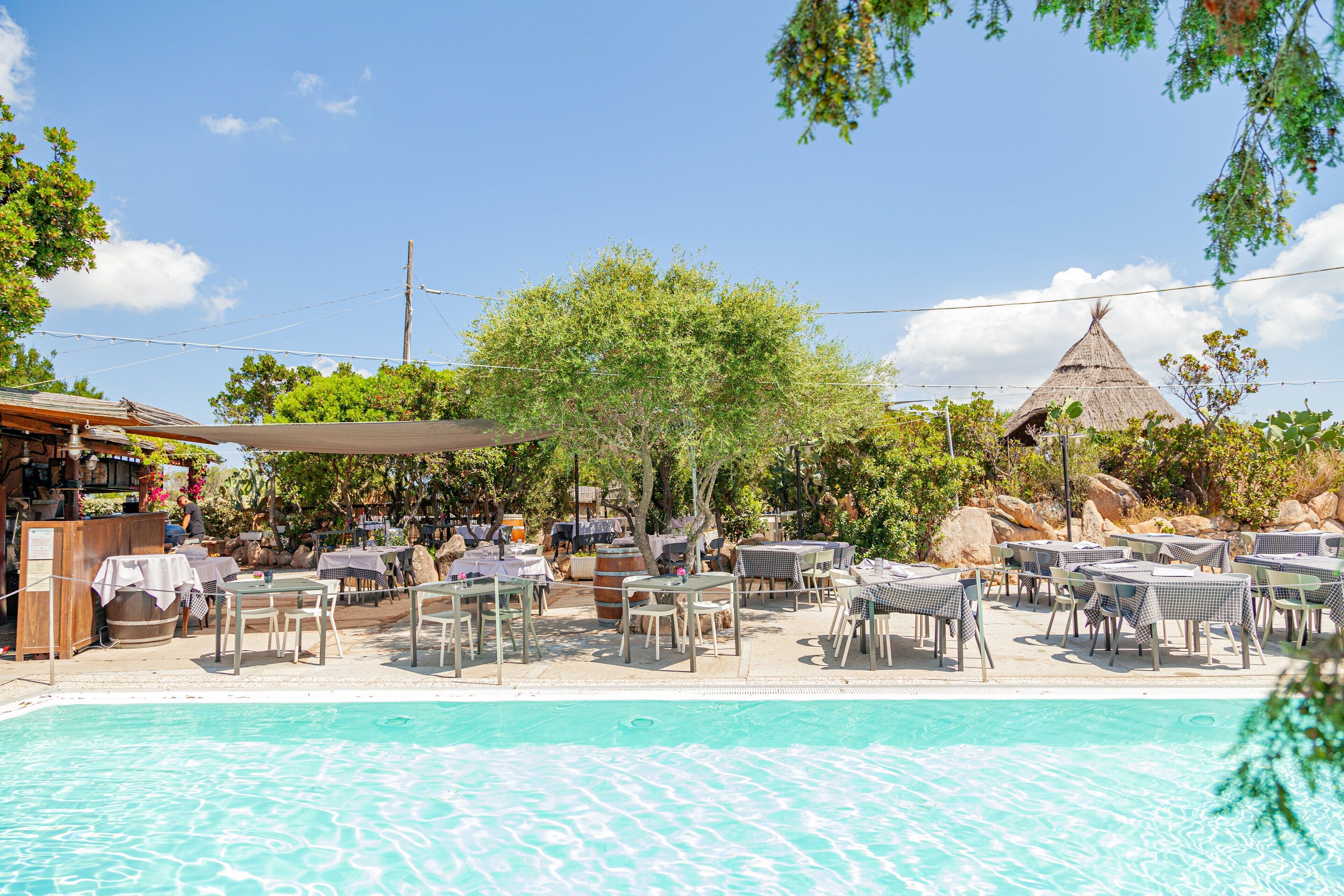 A poolside area with tables and chairs, a bar, and a thatched-roof structure.