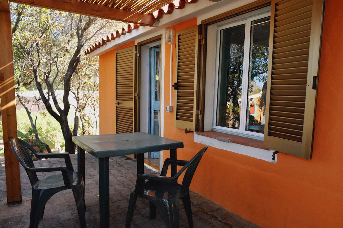 A patio with a table and two chairs, a wooden pergola overhead, and a building with orange walls and shutters.