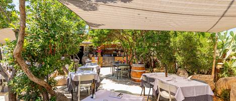 An outdoor dining area with tables covered in white tablecloths, surrounded by greenery and trees.