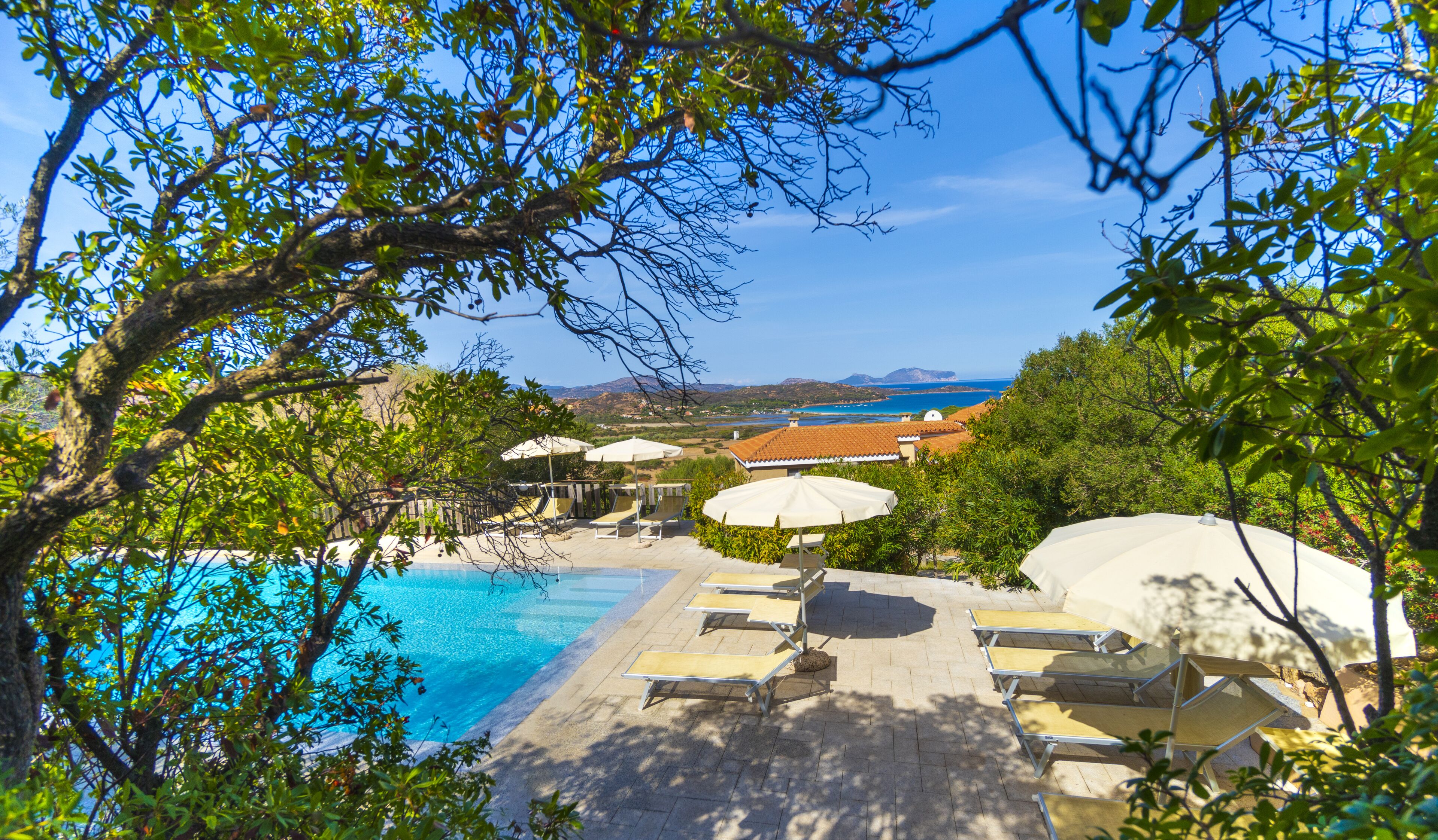 Zona de piscina con sillones de descanso y sombrillas, rodeada de árboles y con vistas a un paisaje pintoresco.