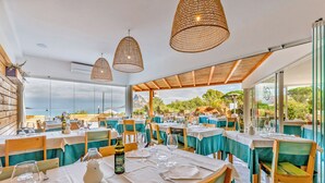 Un restaurante con mesas preparadas para comer, grandes lámparas colgantes de mimbre y vista al mar.