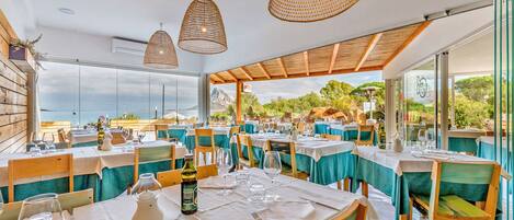 A restaurant with tables set for dining, large hanging wicker lamps, and a view of the sea.