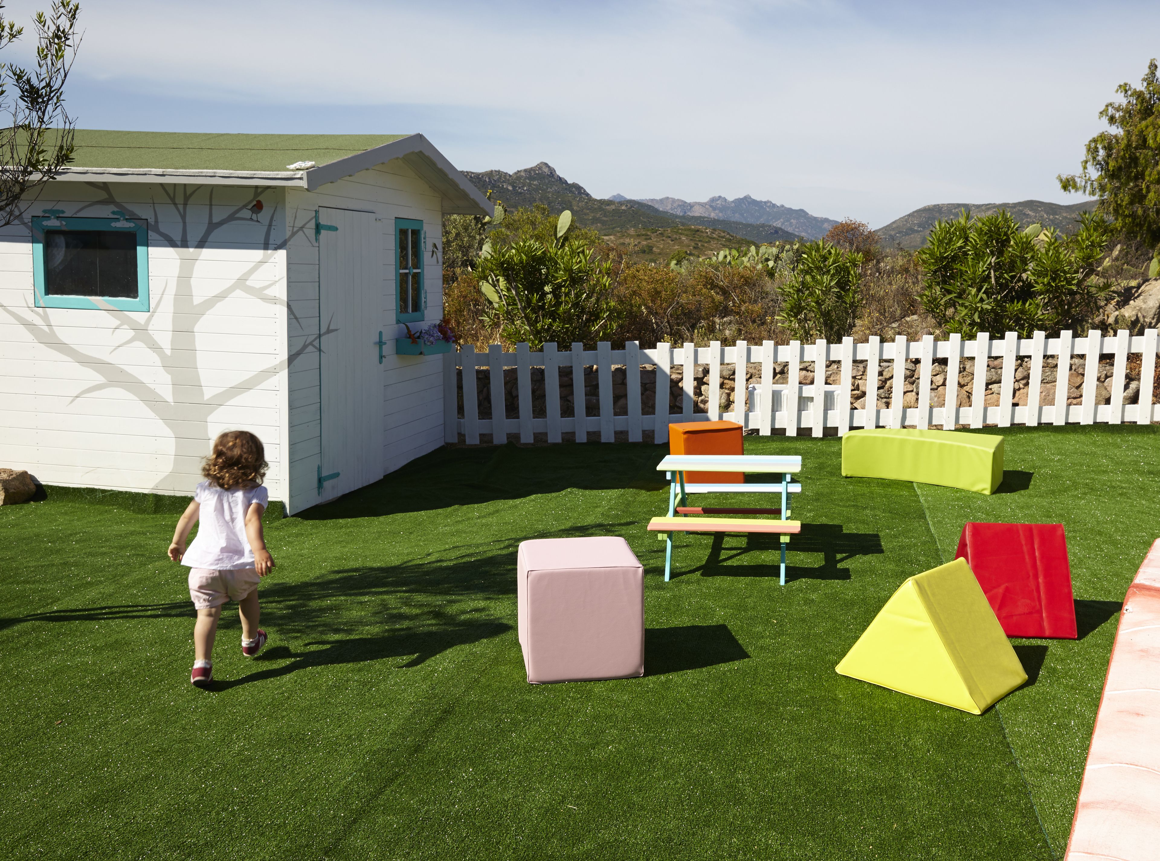 A child playing in a backyard with a white playhouse, green artificial grass, and colorful play structures.