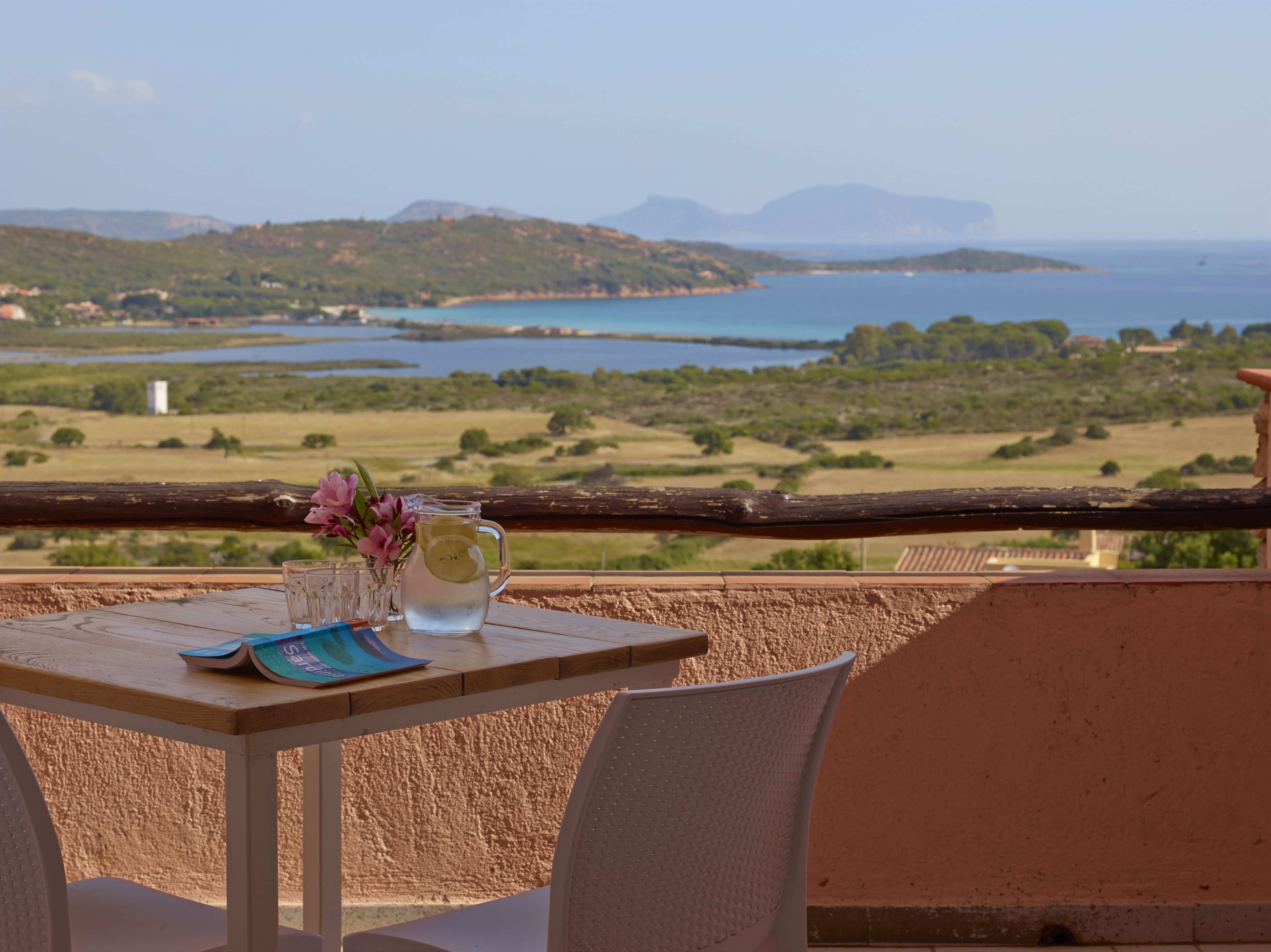 A table with a pitcher of water, a glass, and a menu on a balcony overlooking a coastal landscape with hills and a body of water.