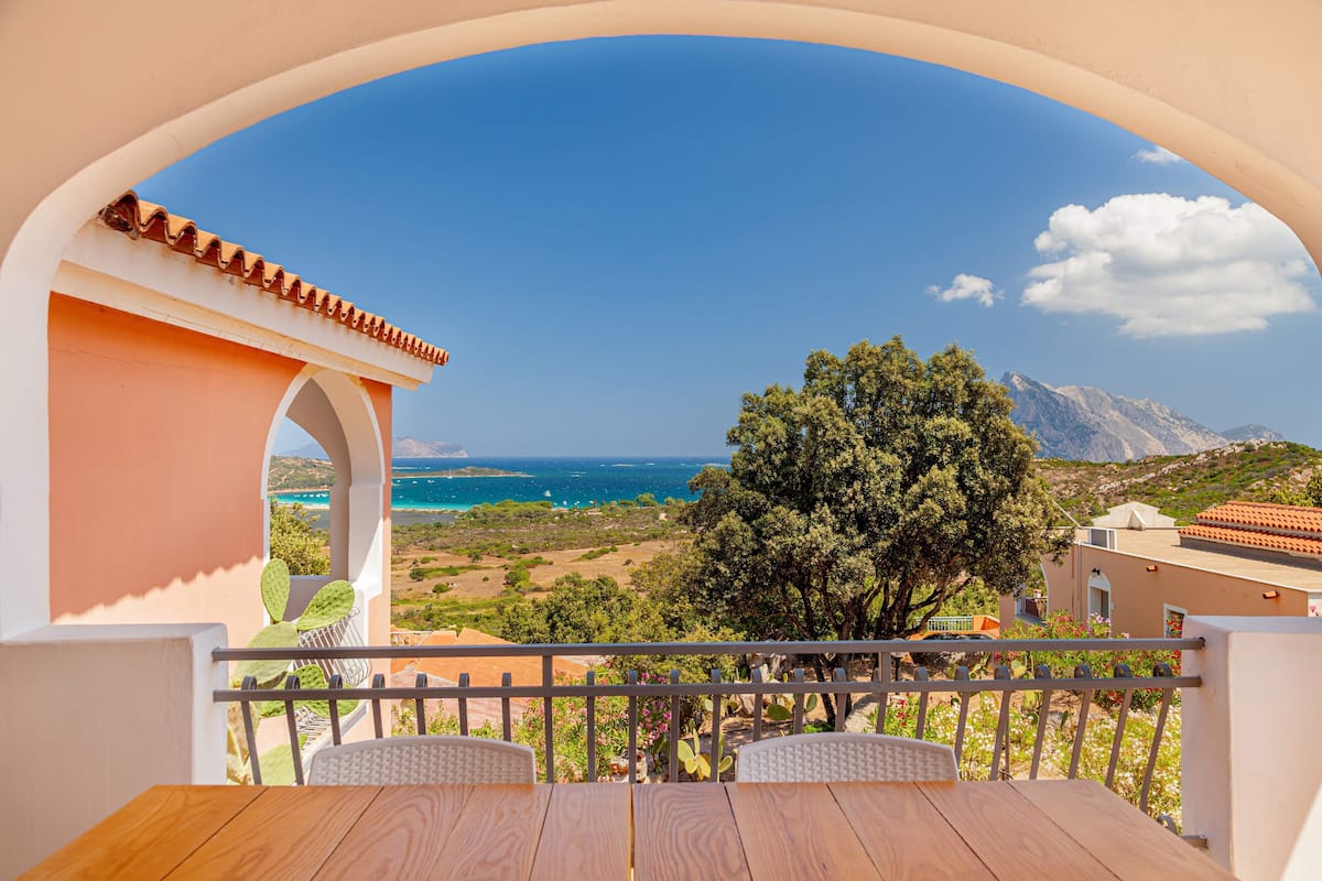 A balcony with a wooden table and chairs, overlooking a coastal landscape with a clear sky and distant mountains.
