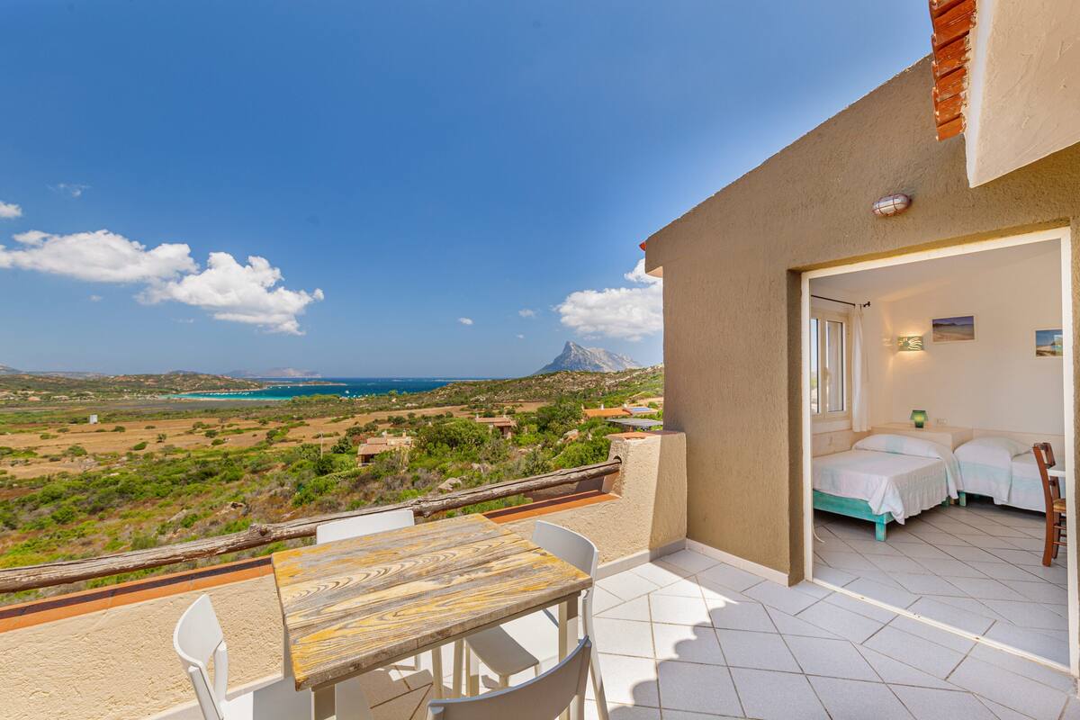 A balcony with a wooden table and chairs, a bed, and a view of a mountainous landscape and sea.