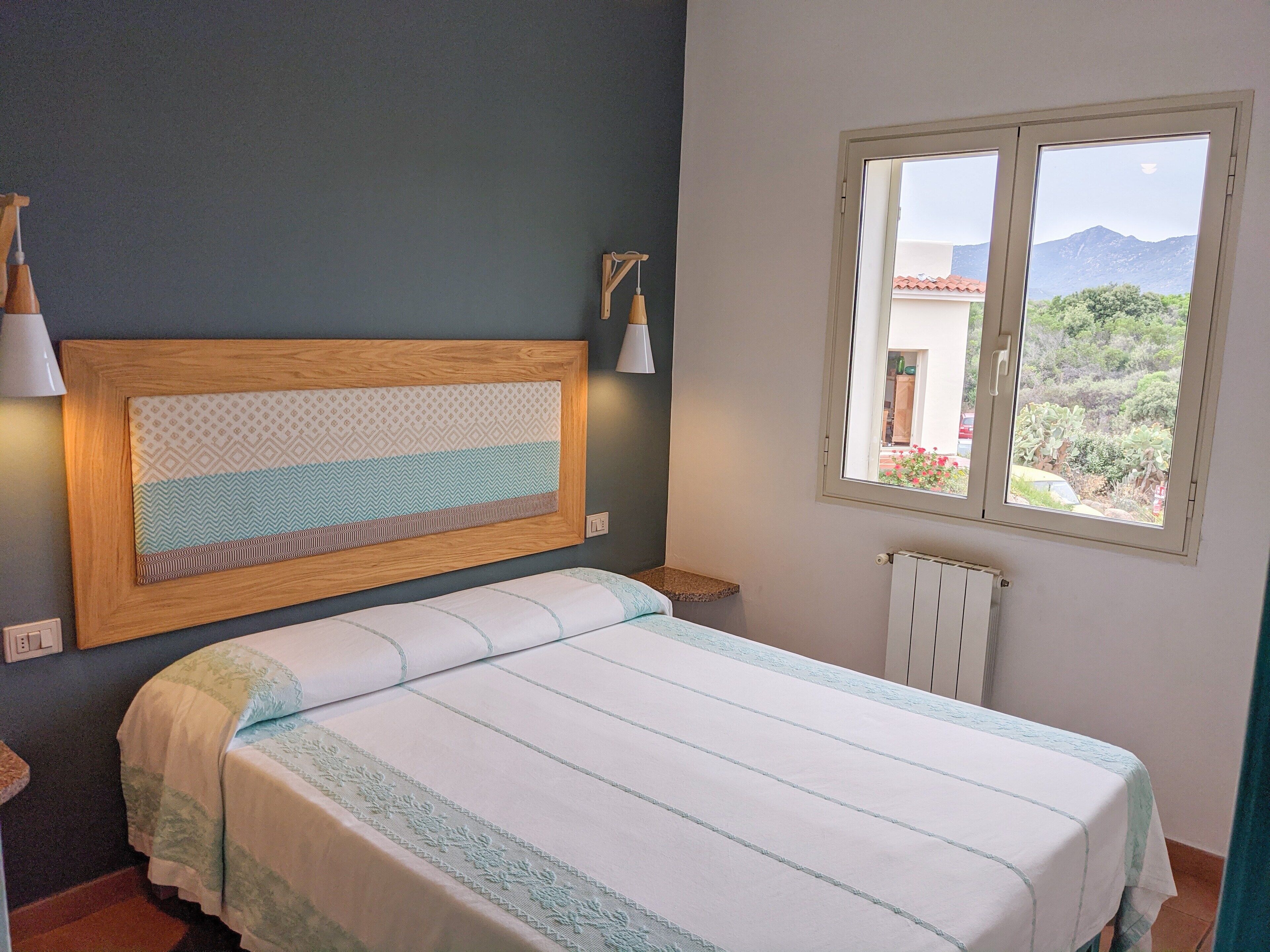 A bedroom with a bed, a window with a view of greenery and mountains, and a wall-mounted light fixture.