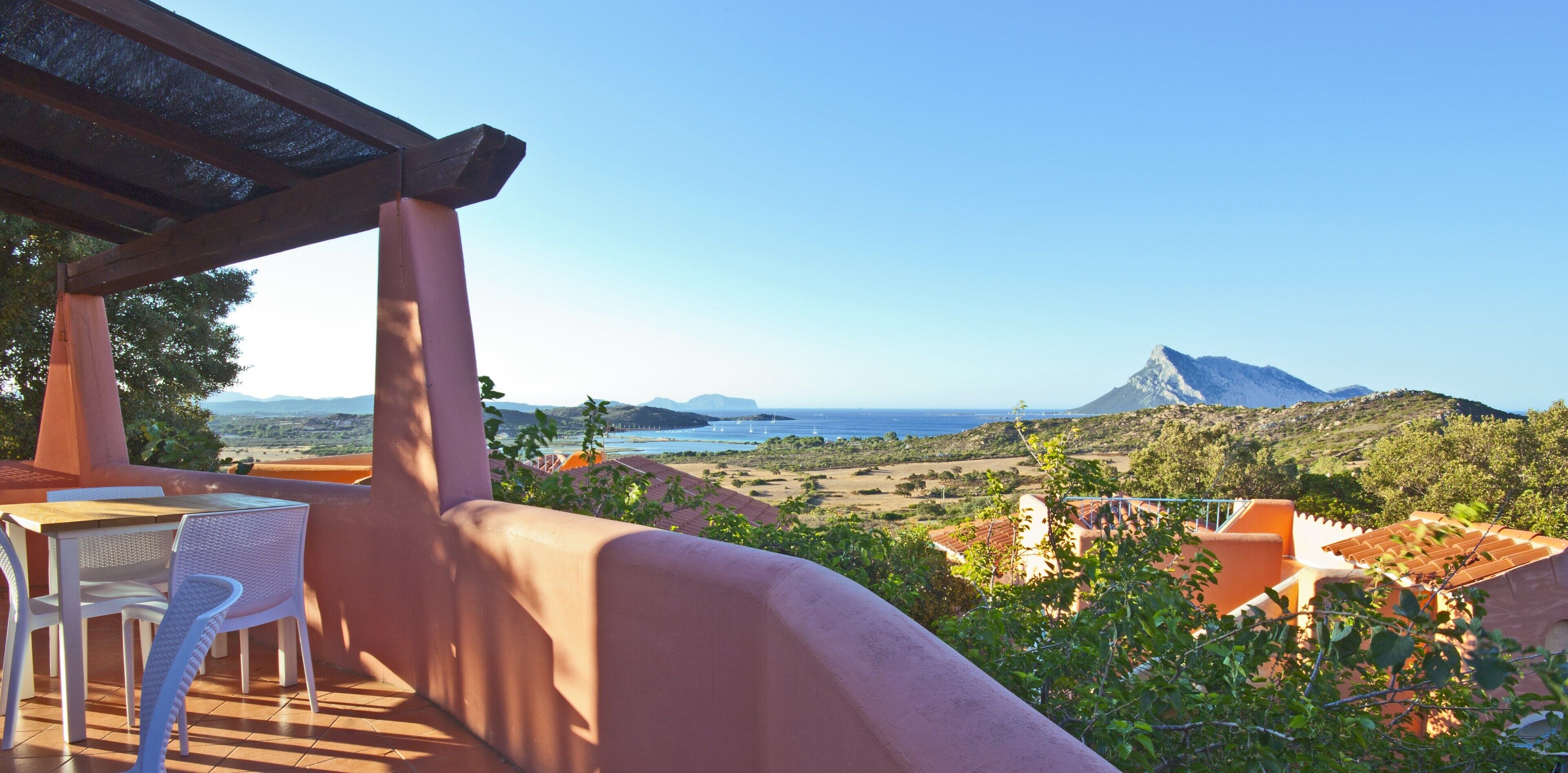 A terrace with a table and chairs, overlooking a coastal landscape with mountains and a clear sky.