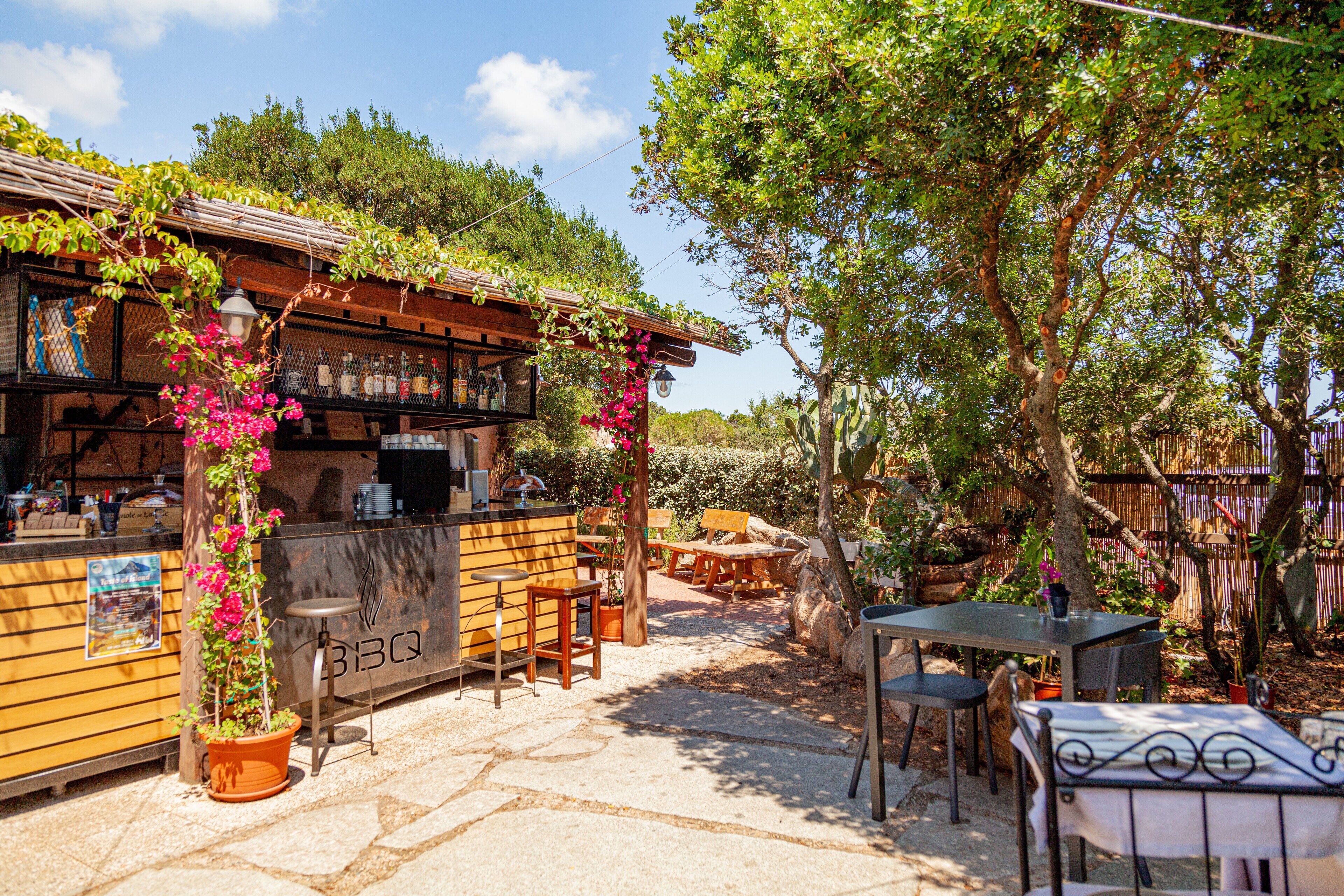 An outdoor seating area with wooden tables and chairs, a bar counter, and potted plants.