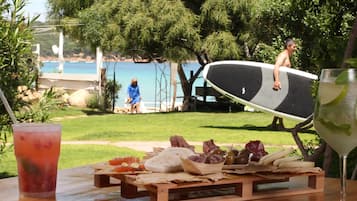 A picnic setup with a table, food, and a drink, overlooking a beach and a pool.