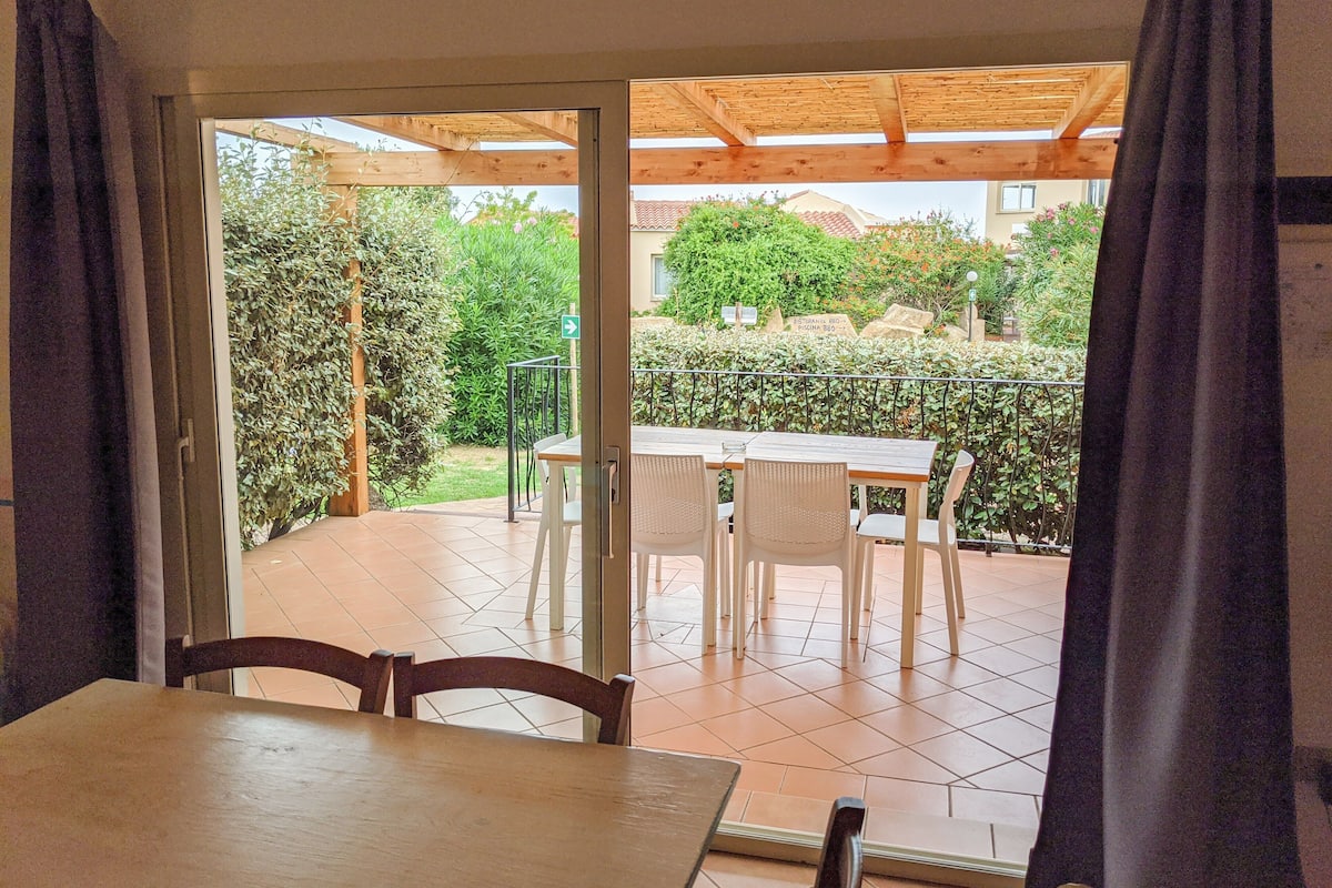 An open patio with a table and chairs, a wooden pergola, and a view of a garden and building in the background.