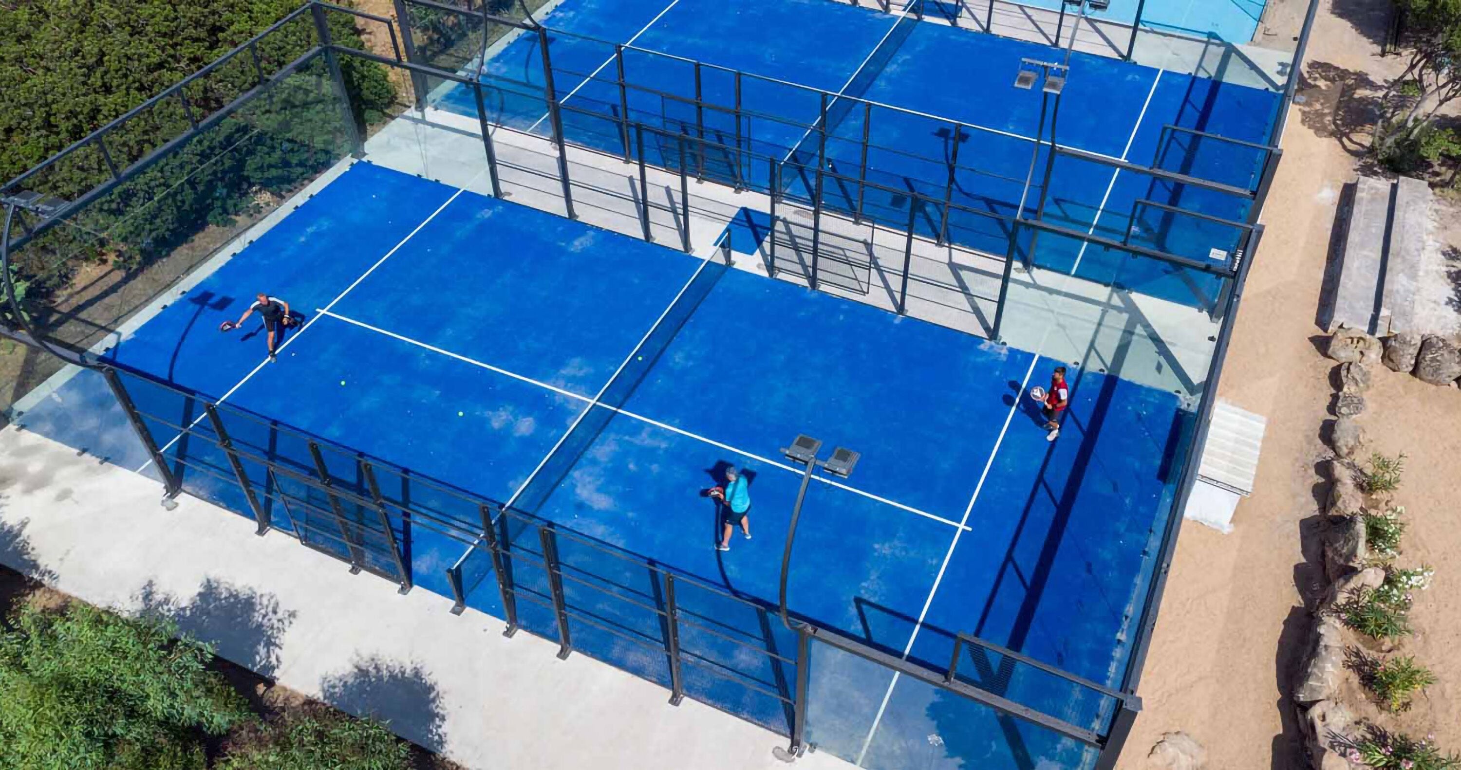 An aerial view of a blue tennis court with a metal fence, surrounded by greenery and a sandy area with rocks.