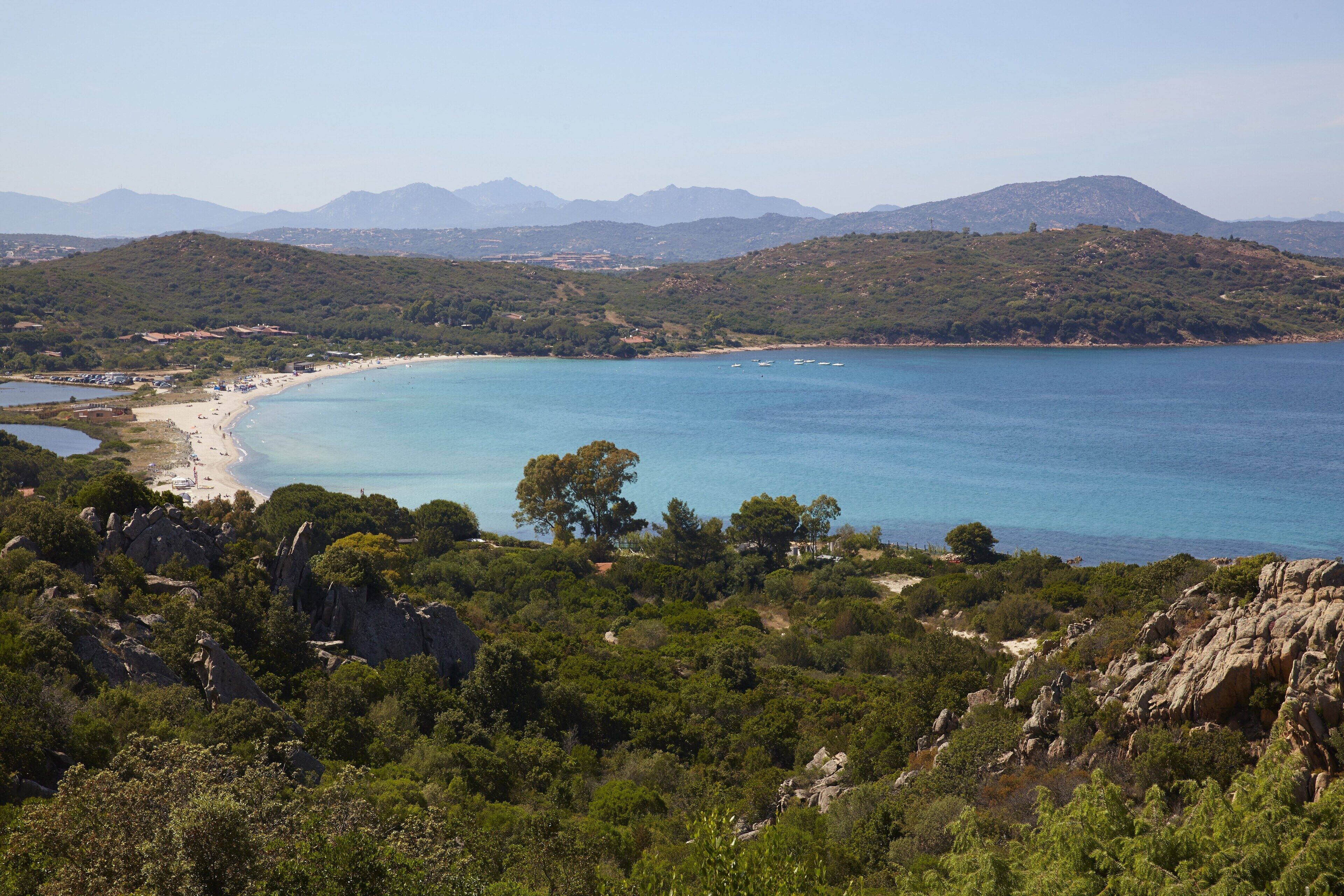 A coastal landscape with a beach, clear blue sea, and mountainous background.