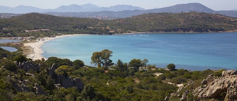 A coastal landscape with a beach, clear blue sea, and mountainous background.