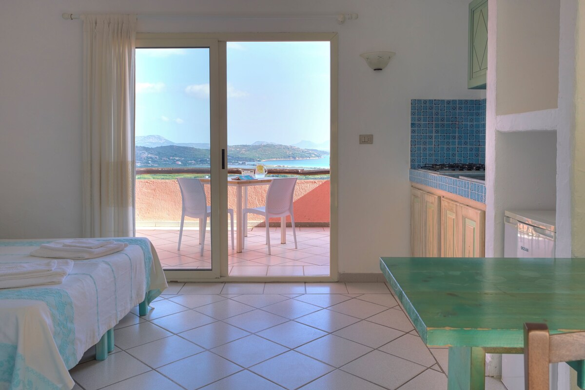 A room with a bed, a table, and a view of a beach and mountains.