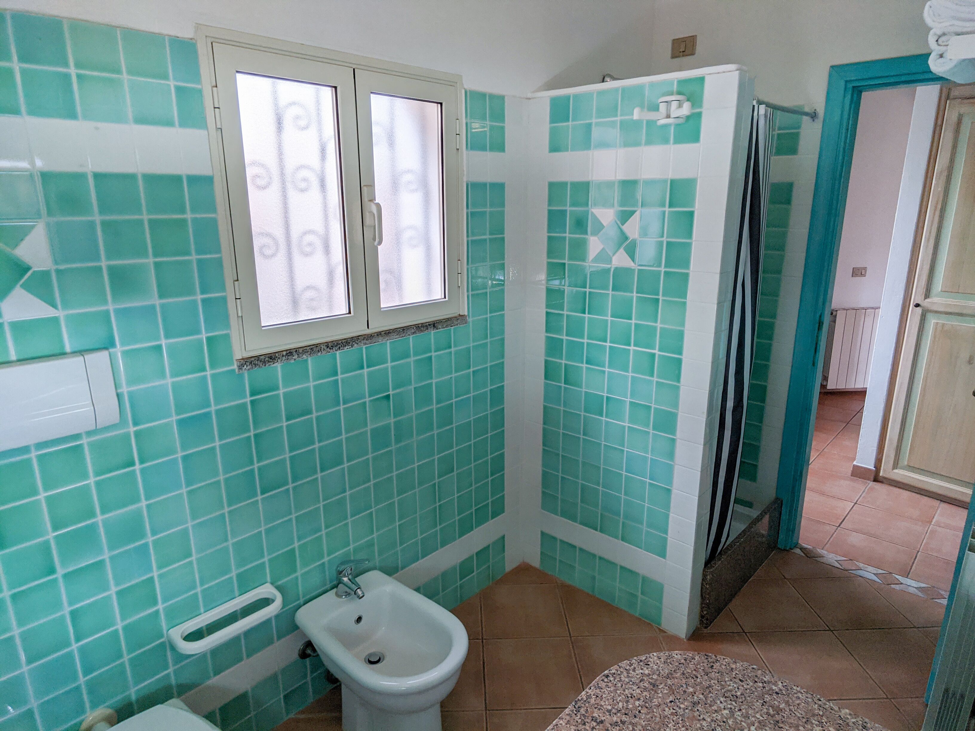 A bathroom with turquoise tiles, a bidet, and a window with frosted glass.