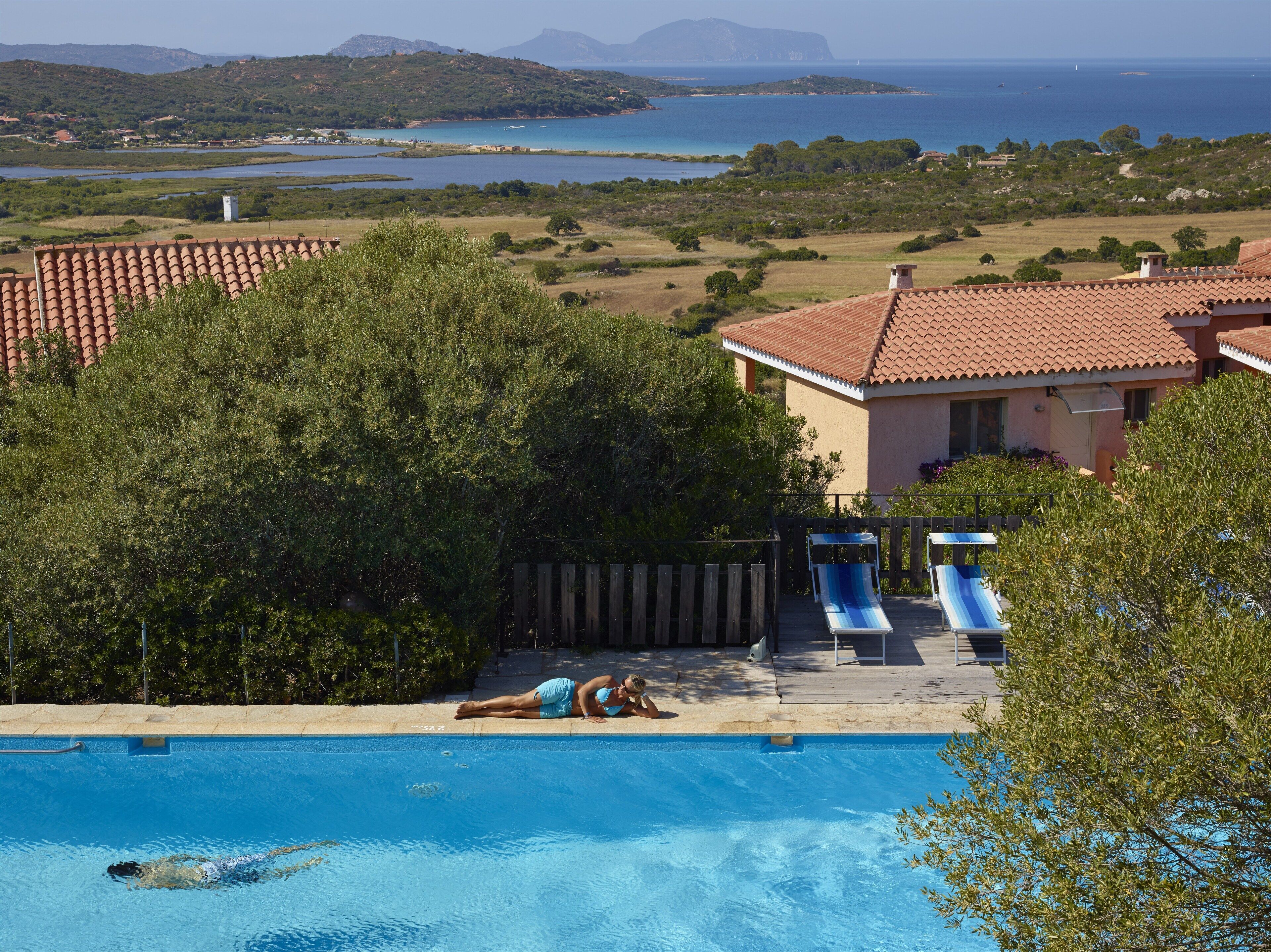 A swimming pool with a person lying on the edge, overlooking a coastal landscape with hills and a body of water.
