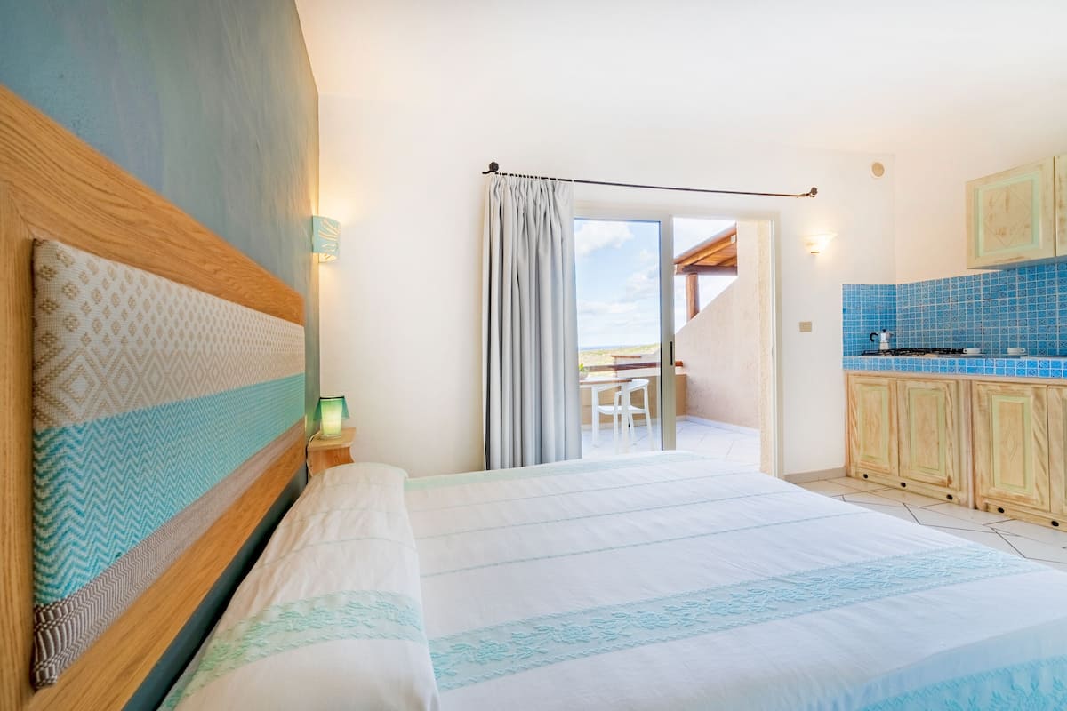 A bedroom with a large bed, a view to a balcony, and a kitchenette with blue tiles.