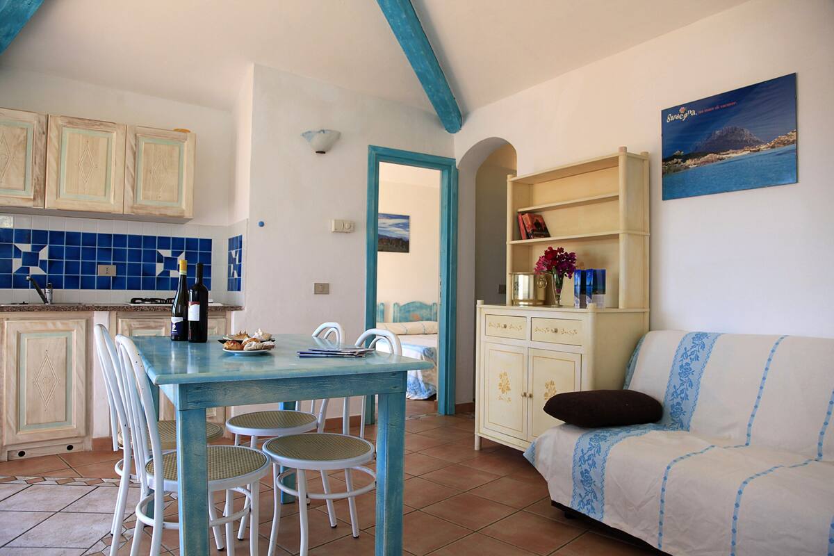 A dining area with a table and chairs, a kitchen with blue tiles, and a sofa with a decorative pillow.