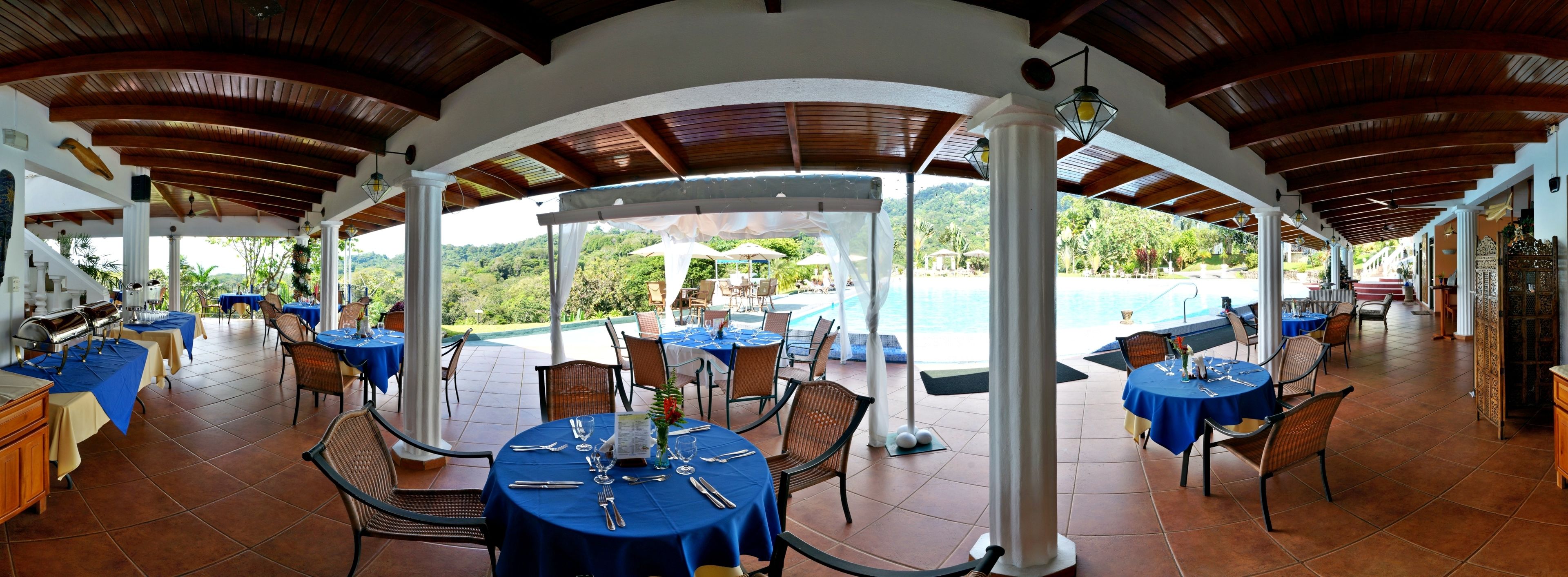 Un área de comedor al aire libre techada con mesas dispuestas para una comida, con vista a una piscina y abundante vegetación.