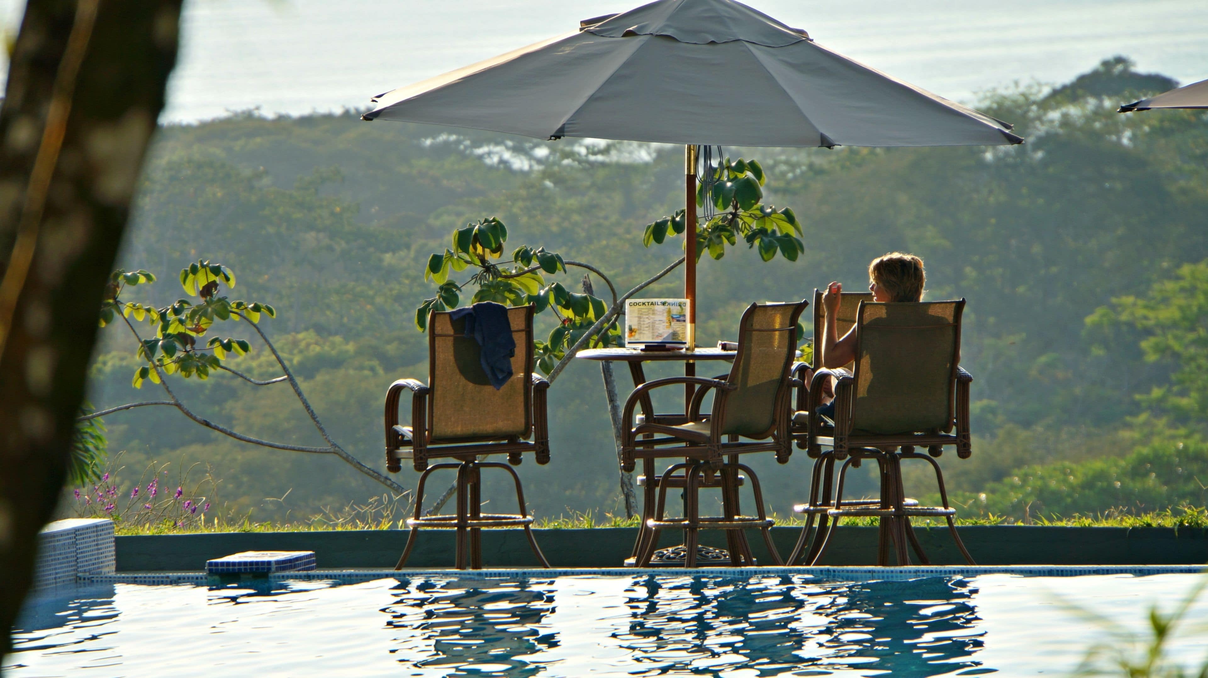 A poolside setting with a table and chairs under an umbrella, surrounded by lush greenery.
