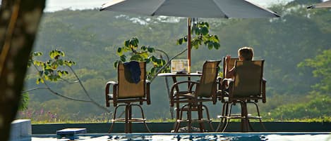 A poolside setting with a table and chairs under an umbrella, surrounded by lush greenery.