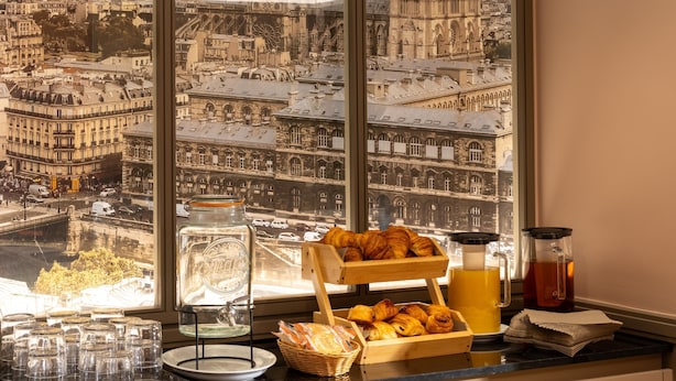 A breakfast setup with croissants, a coffee pot, and a teapot on a table by a window with a city view. - Hotel Paris Bastille (Paris)