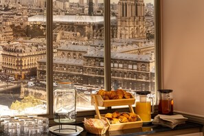 A breakfast setup with croissants, a coffee pot, and a teapot on a table by a window with a city view. - Hotel Paris Bastille (Paris)