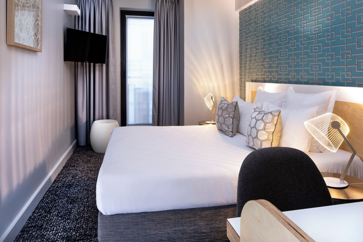 A hotel room with a large bed, a bedside table with a lamp, a wall-mounted TV, and a view of the outdoors.