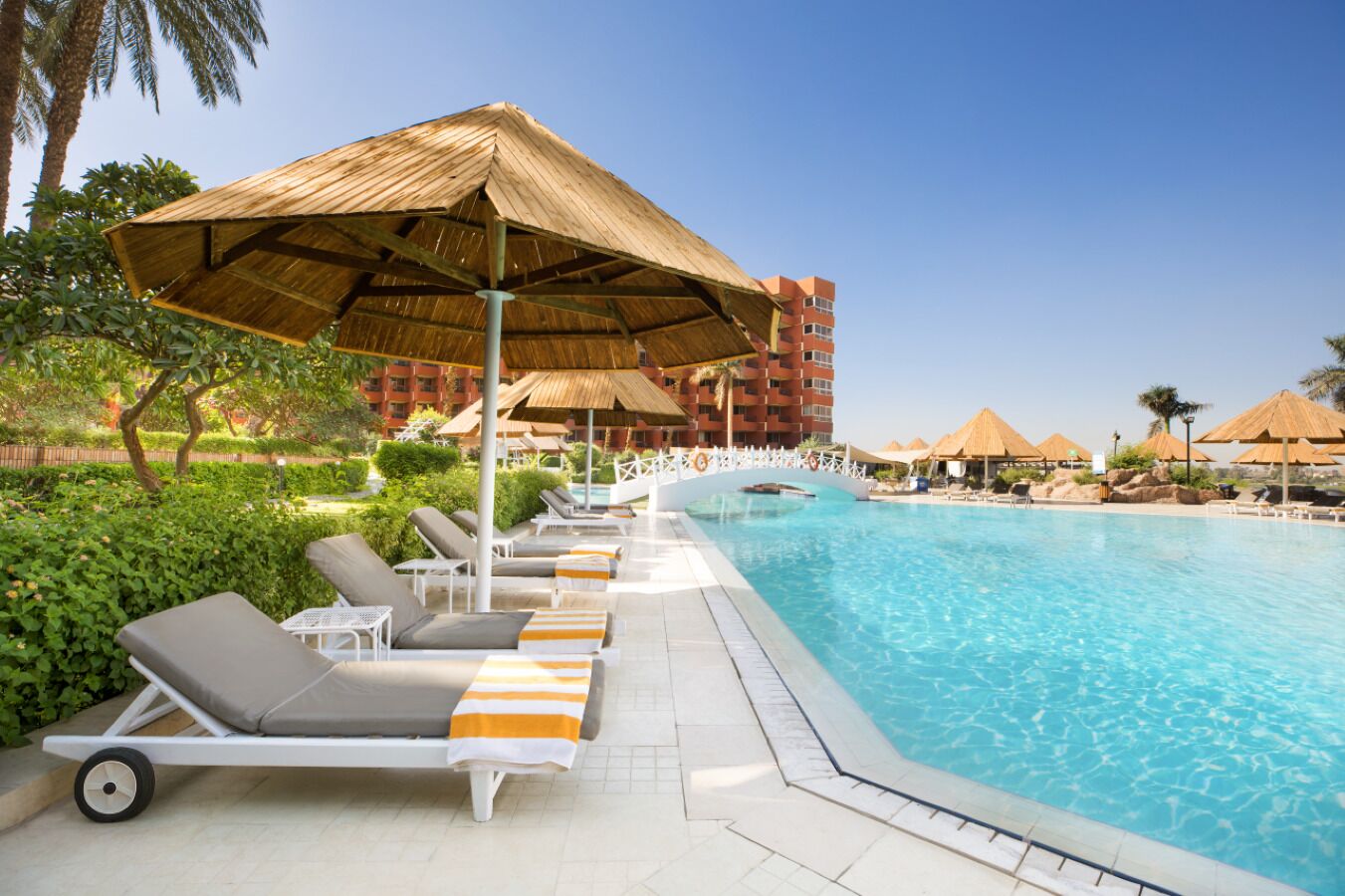A poolside area with sun loungers and thatched umbrellas, surrounded by lush greenery and a clear blue pool.