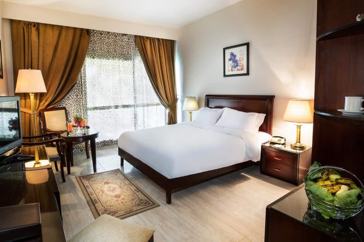 Una habitación de hotel con cama, mesitas de noche, un escritorio con lámpara, televisor, mesa de centro y un cuenco decorativo.