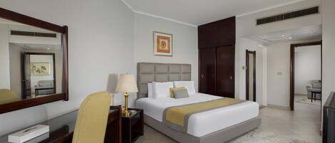 A hotel room with a large bed, a bedside table with a lamp, a chair, a desk with a computer, a TV, and a wardrobe.