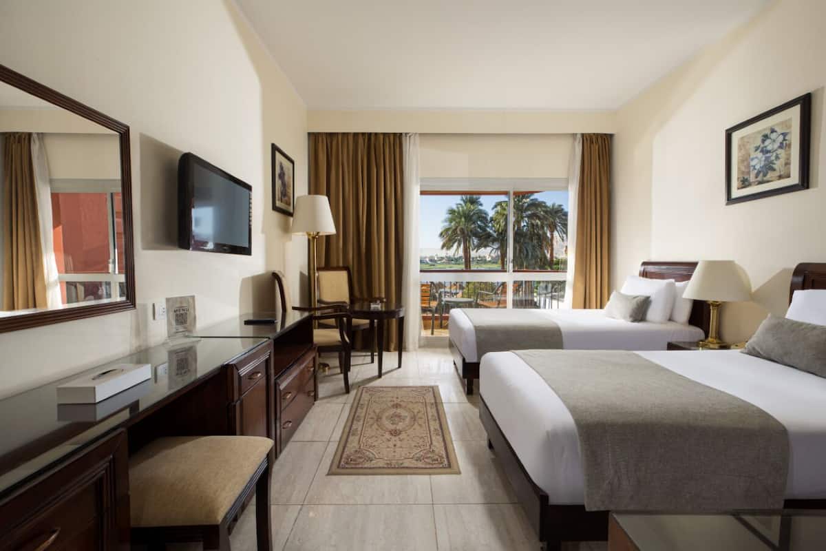 Habitación de hotel con dos camas, un escritorio, una silla, un televisor y un balcón con vistas a palmeras.