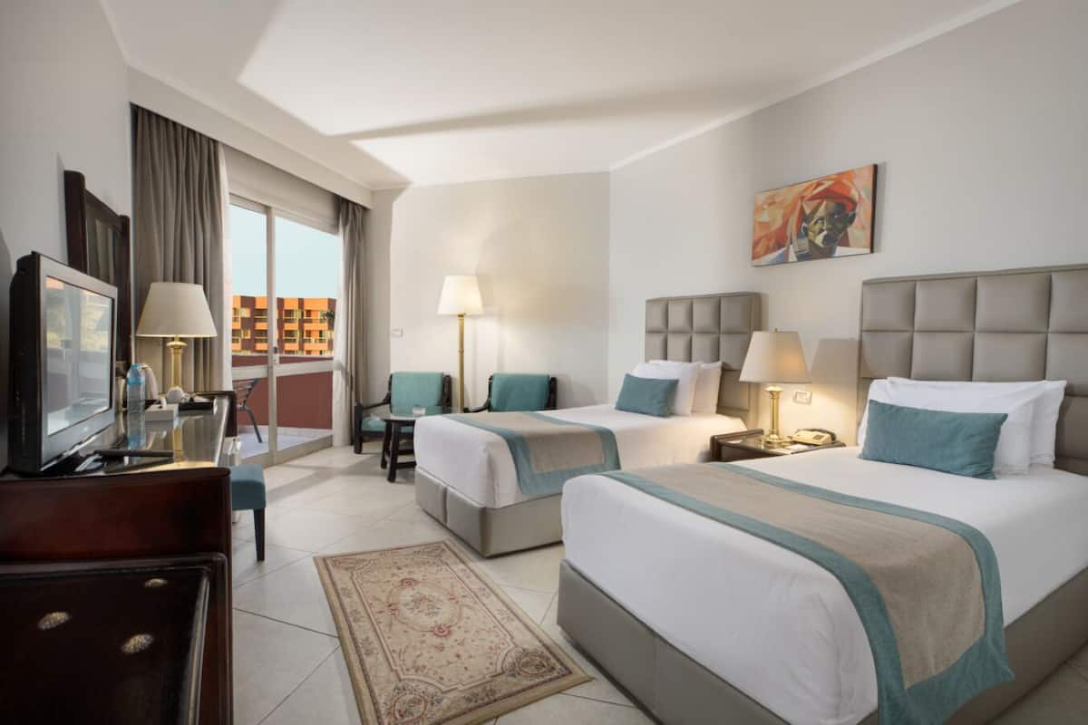 Habitación de hotel con dos camas, un escritorio con televisor, una silla, una ventana con cortinas y un cuadro en la pared.