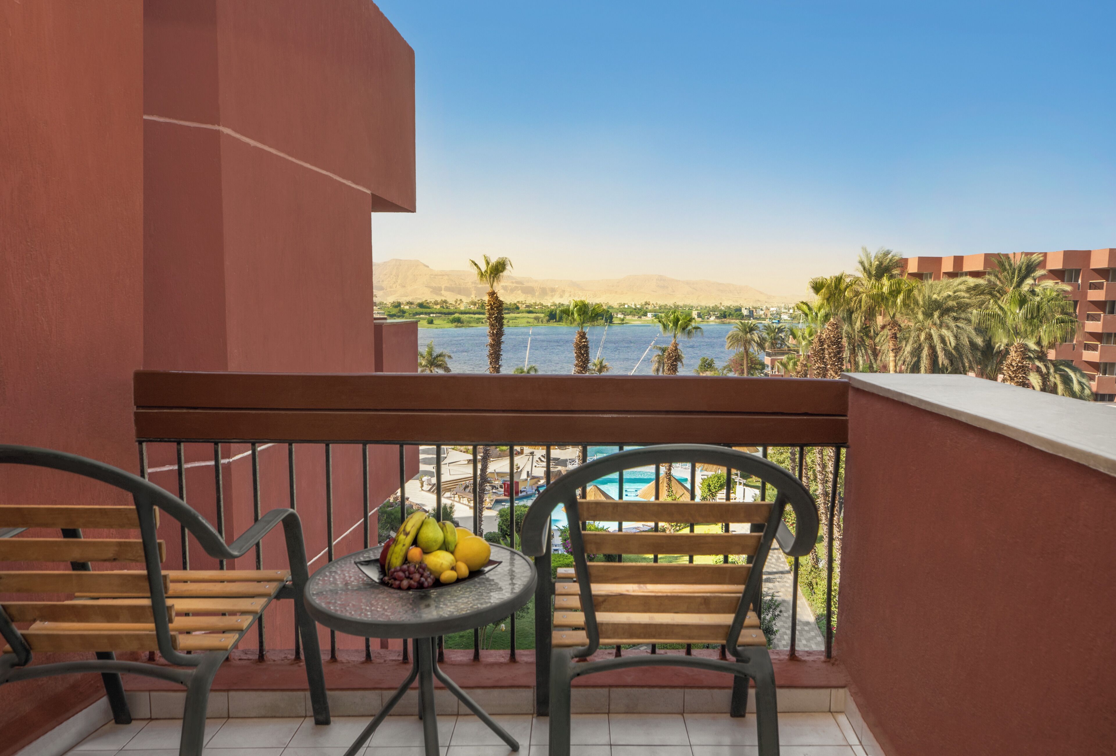 A balcony with two chairs and a small table with fruit, overlooking a body of water and palm trees.