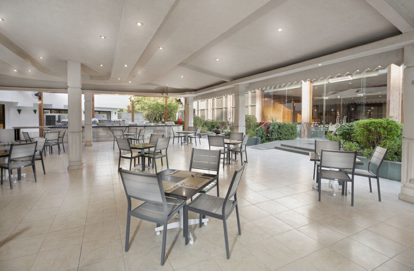 Un amplio comedor interior con varias mesas y sillas, una zona de cocina al fondo, y vistas al exterior a través de grandes ventanales.